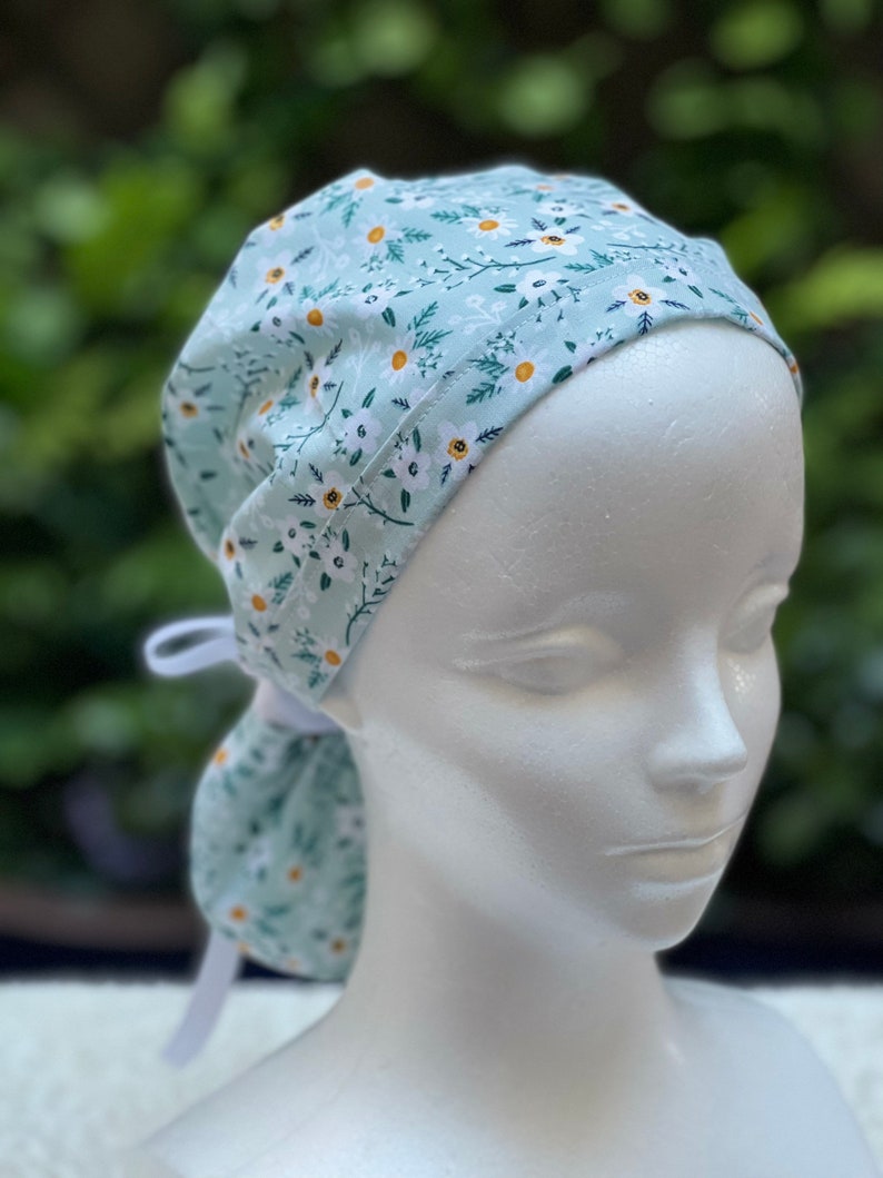 K&ouml;nnte beinhalten: Eine hellblaue OP-M&uuml;tze mit wei&szlig;em Blumenmuster. Die M&uuml;tze hat ein wei&szlig;es Band, das am Hinterkopf gebunden werden kann.