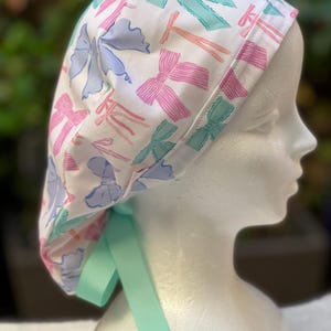 May include: A white surgical scrub hat with a colourful bow print. The hat has a light blue, pink, and green bow pattern. The hat has a light green tie.