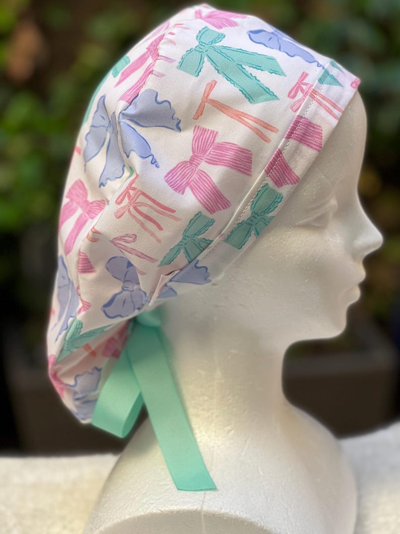 May include: A white surgical scrub hat with a colourful bow print. The hat has a light blue, pink, and green bow pattern. The hat has a light green tie.