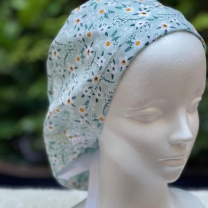 K&ouml;nnte beinhalten: Eine hellblaue OP-M&uuml;tze mit einem wei&szlig;en und gelben Blumenmuster. Die M&uuml;tze hat wei&szlig;e B&auml;nder.