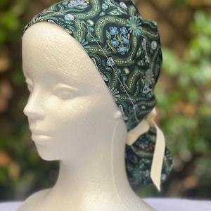 May include: A dark green patterned scrub cap with a floral design in shades of blue and green. The cap is tied in the back with a cream-colored ribbon. The cap is displayed on a white mannequin head.