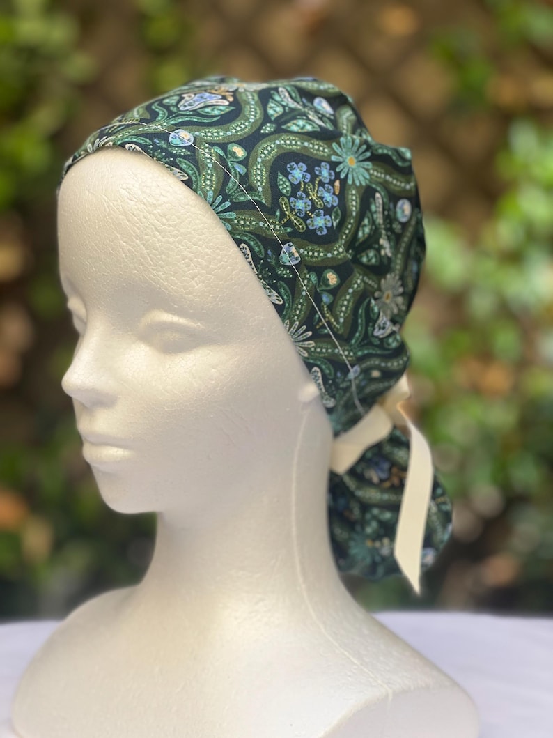 May include: A dark green patterned scrub cap with a floral design in shades of blue and green. The cap is tied in the back with a cream-colored ribbon. The cap is displayed on a white mannequin head.