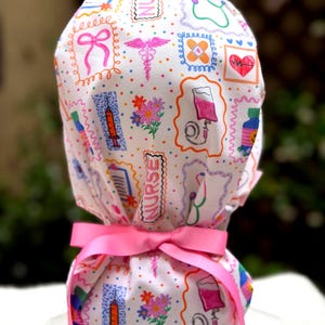 May include: A white medical scrub cap with a pink ribbon tie. The cap is patterned with colorful medical-themed illustrations, including syringes, stethoscopes, and flowers. The design features a repeating pattern of medical symbols and the word "NURSE".