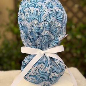 May include: A blue and white patterned scrub cap with a wave design. The cap is tied with a white ribbon bow. The fabric features a repeating pattern of stylized ocean waves in shades of blue and white.