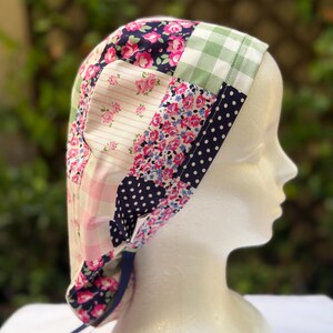 May include: A patchwork fabric cap with a variety of floral, polka dot, and gingham patterns in pink, navy, and green. The cap has a dark blue ribbon tie and is displayed on a mannequin head.