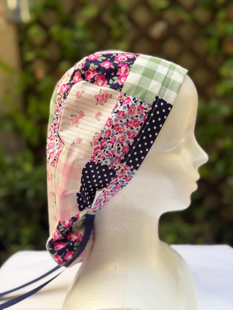 May include: A patchwork fabric cap with a variety of floral, polka dot, and gingham patterns in pink, navy, and green. The cap has a dark blue ribbon tie and is displayed on a mannequin head.