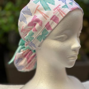 May include: A white surgical scrub hat with a colourful bow pattern. The hat has a tie that can be tied at the back of the neck.