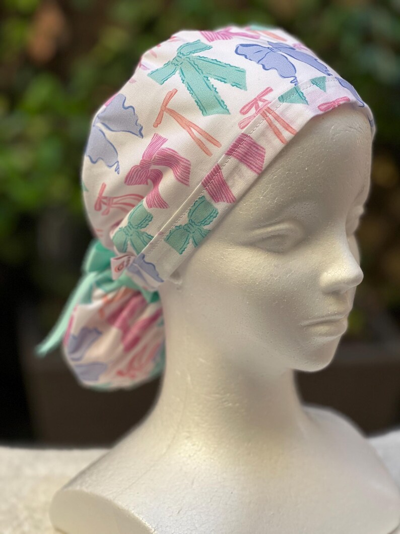 May include: A white surgical scrub hat with a colourful bow pattern. The hat has a tie that can be tied at the back of the neck.