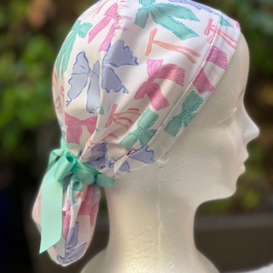 May include: A white surgical scrub hat with a colourful bow pattern. The hat has a tie at the back.