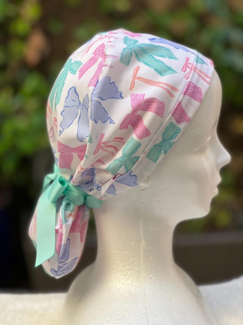 May include: A white surgical scrub hat with a colourful bow pattern. The hat has a tie at the back.
