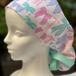 May include: A white surgical scrub hat with a colourful bow pattern. The hat has a light blue ribbon tie.