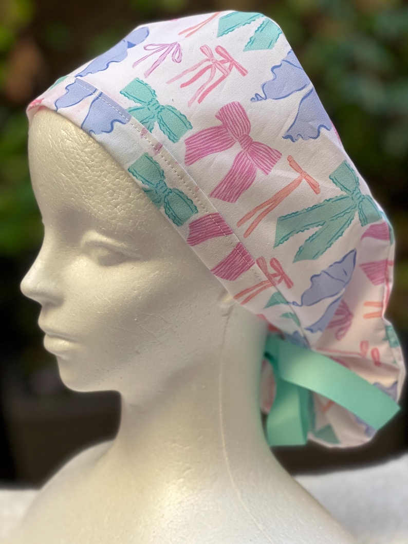 May include: A white surgical scrub hat with a colourful bow pattern. The hat has a light blue ribbon tie.