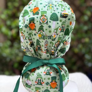 May include: A light green scrub cap with a repeating pattern of gnomes, shamrocks, and mushrooms. The cap is tied with a dark green ribbon. The word "LUCK" is visible. Designed for medical professionals.