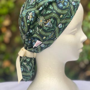 May include: A dark green patterned scrub cap with a floral design in shades of blue, white, and green. The cap is tied at the back with a cream-colored ribbon. The cap is on a mannequin head.