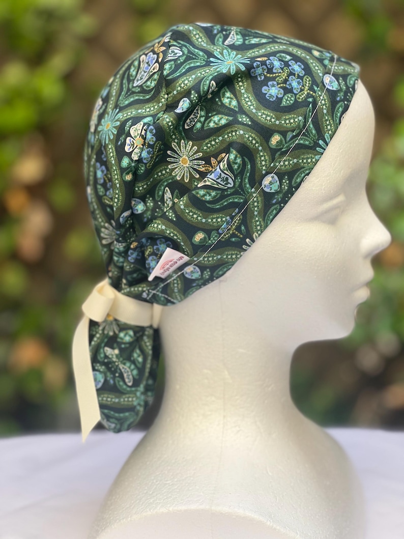 May include: A dark green patterned scrub cap with a floral design in shades of blue, white, and green. The cap is tied at the back with a cream-colored ribbon. The cap is on a mannequin head.