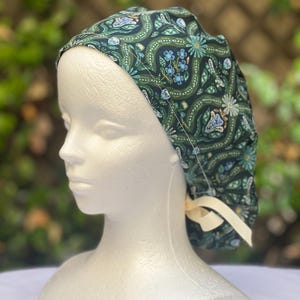 May include: A dark green patterned scrub cap featuring a floral and butterfly design in shades of teal, blue, and cream. The cap is tied with a cream-colored ribbon. The cap is on a white mannequin head.