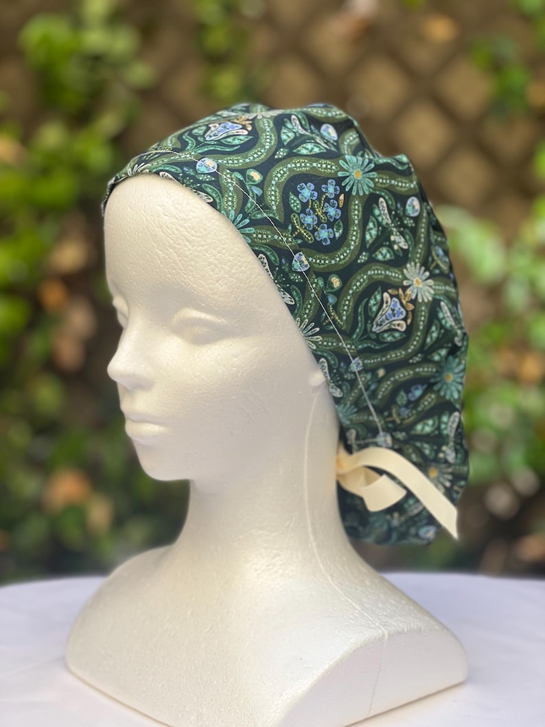May include: A dark green patterned scrub cap featuring a floral and butterfly design in shades of teal, blue, and cream. The cap is tied with a cream-colored ribbon. The cap is on a white mannequin head.