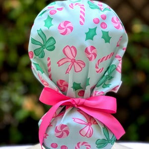 May include: A light blue scrub cap with a festive pattern of pink and green candy canes, bows, and swirls. The cap is tied with a bright pink ribbon. The background is blurred green foliage.
