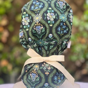 May include: A dark blue patterned scrub cap with a floral design in shades of green, blue, and cream. The cap is tied with a cream-colored ribbon. The fabric features a repeating pattern of stylized flowers and leaves.