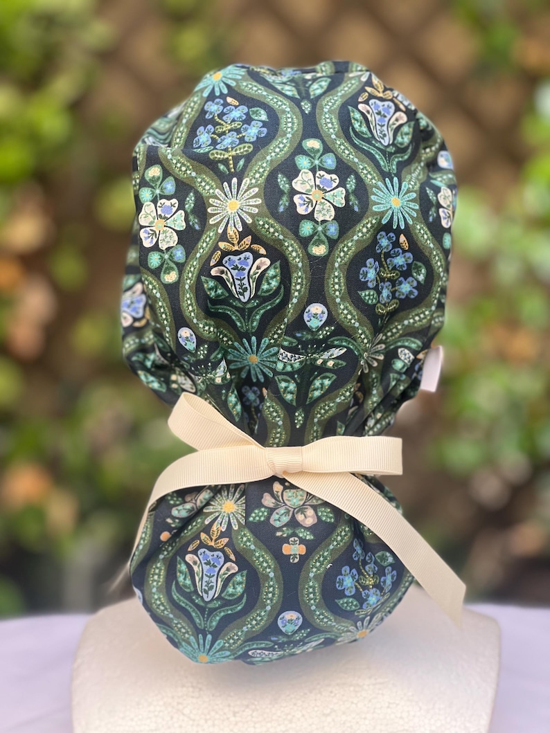 May include: A dark blue patterned scrub cap with a floral design in shades of green, blue, and cream. The cap is tied with a cream-colored ribbon. The fabric features a repeating pattern of stylized flowers and leaves.