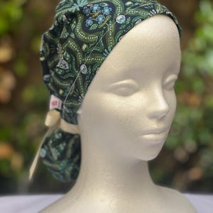May include: A dark green patterned scrub cap with a floral design in shades of green and blue. The cap is tied in the back with a cream-colored ribbon. The cap is on a mannequin head.