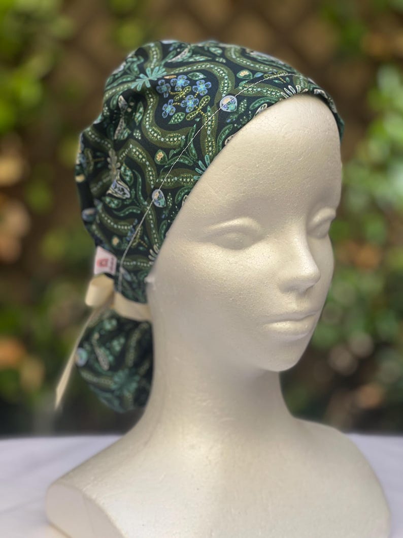 May include: A dark green patterned scrub cap with a floral design in shades of green and blue. The cap is tied in the back with a cream-colored ribbon. The cap is on a mannequin head.
