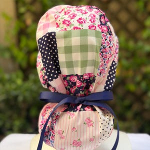 May include: A patchwork scrub cap with floral, polka dot, and gingham patterns in pink, navy, and green. It is tied with a navy blue ribbon.