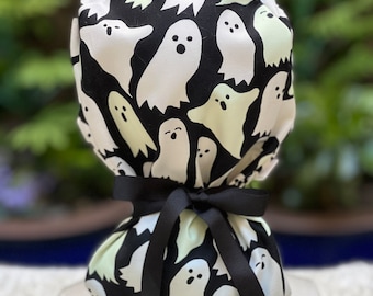 May include: A black and white Halloween-themed scrub cap featuring a pattern of white ghost figures on a black background. The cap is tied with a black ribbon. The ghosts have simple, friendly faces.