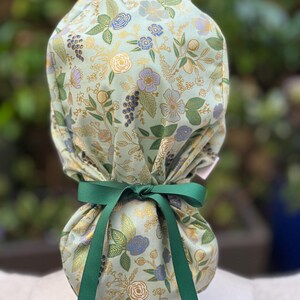 May include: A light green scrub hat with a floral print featuring gold, blue, and purple flowers. The hat has a green ribbon tied around it.
