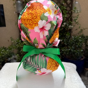 May include: A scrub cap featuring a vibrant pink background with a tropical pattern of yellow pineapples, white and pink hibiscus flowers, and green palm leaves. The cap is secured with a bright green ribbon tied into a bow at the back.