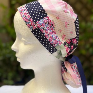 May include: A patchwork scrub cap with a variety of patterns, including navy blue polka dots, pink gingham, and floral prints. A navy blue ribbon is tied at the back. The cap is displayed on a white mannequin head.