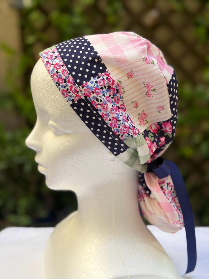 May include: A patchwork scrub cap with a variety of patterns, including navy blue polka dots, pink gingham, and floral prints. A navy blue ribbon is tied at the back. The cap is displayed on a white mannequin head.