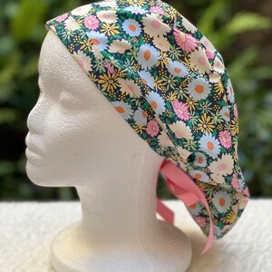 May include: A surgical scrub hat with a floral print featuring pink, yellow, and white daisies on a dark blue background. The hat has a pink ribbon tie.