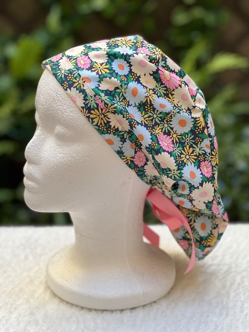 May include: A surgical scrub hat with a floral print featuring pink, yellow, and white daisies on a dark blue background. The hat has a pink ribbon tie.