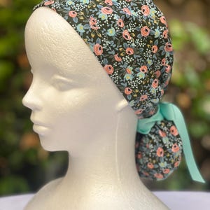 May include: A floral print scrub cap featuring a dark background with pink, blue, and white flowers. The cap is tied in the back with a light teal ribbon. The cap is worn on a white mannequin head.