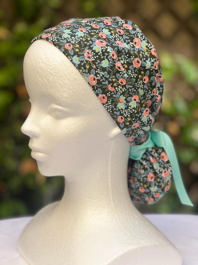 May include: A floral print scrub cap featuring a dark background with pink, blue, and white flowers. The cap is tied in the back with a light teal ribbon. The cap is worn on a white mannequin head.