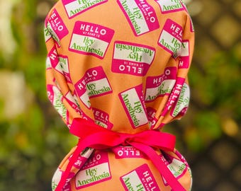 Può includere: Un cappello chirurgico con un motivo ripetuto di etichette rosa e bianche "Hello, Hey Anesthesia" su uno sfondo arancione brillante. Il cappello è legato con un nastro rosa vibrante.