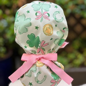 May include: A white scrub cap featuring a St. Patrick's Day theme. The cap is patterned with green shamrocks, gold coins, and horseshoes, accented with pink bows and stars. A pink ribbon is tied around the cap.
