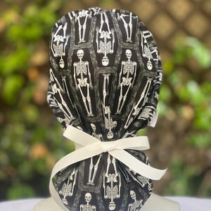 May include: A black and white patterned scrub cap featuring a repeating skeleton design. The cap is tied with a white ribbon bow. The background is blurred, suggesting an outdoor setting. This cap is suitable for medical professionals or Halloween.