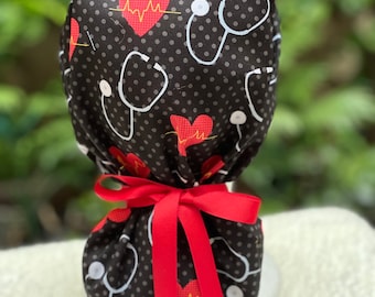 May include: A black scrub cap with a pattern of white stethoscopes, red hearts with a heartbeat design, and small gray polka dots. The cap is tied with a bright red ribbon.