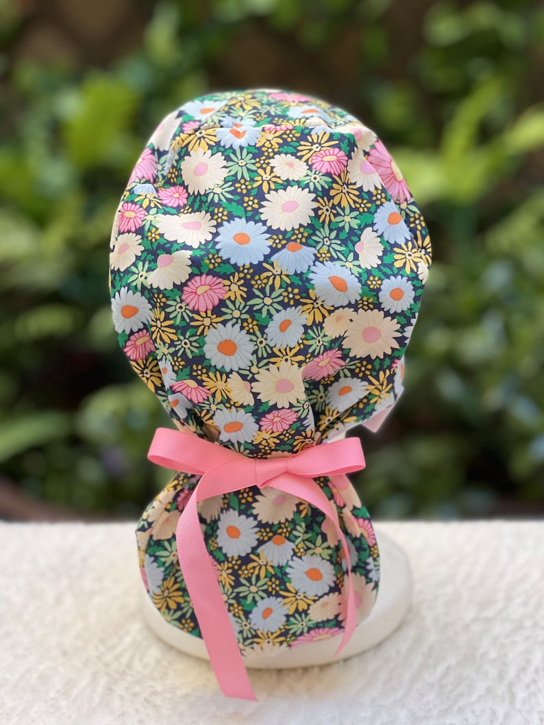 May include: A floral print surgical scrub hat with a pink ribbon tied around the bottom. The hat is made of a soft, comfortable fabric and features a variety of colorful flowers.