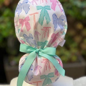 May include: A white scrub hat with a pattern of colourful bows. The hat has a green ribbon tied around it.