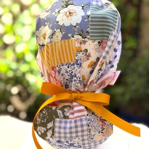 May include: A scrub cap featuring a patchwork design of various floral, striped, and gingham patterns in shades of blue, yellow, white, and green. The fabric is gathered at the back and secured with a solid mustard yellow ribbon tied in a bow.