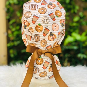 May include: A white and tan patterned scrub cap featuring a repeating design of pumpkins, donuts, coffee cups, and turkeys. The cap is tied with a brown ribbon. The background is blurred, with green foliage.