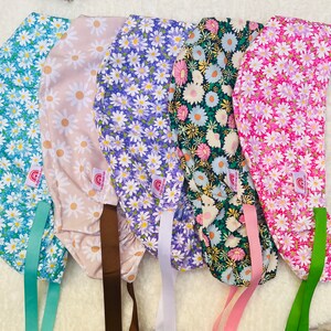 May include: Five colorful scrub hats with floral patterns. The hats are made of fabric and have ties to secure them to the head. The hats are in the following colors: blue, pink, purple, beige, and green.