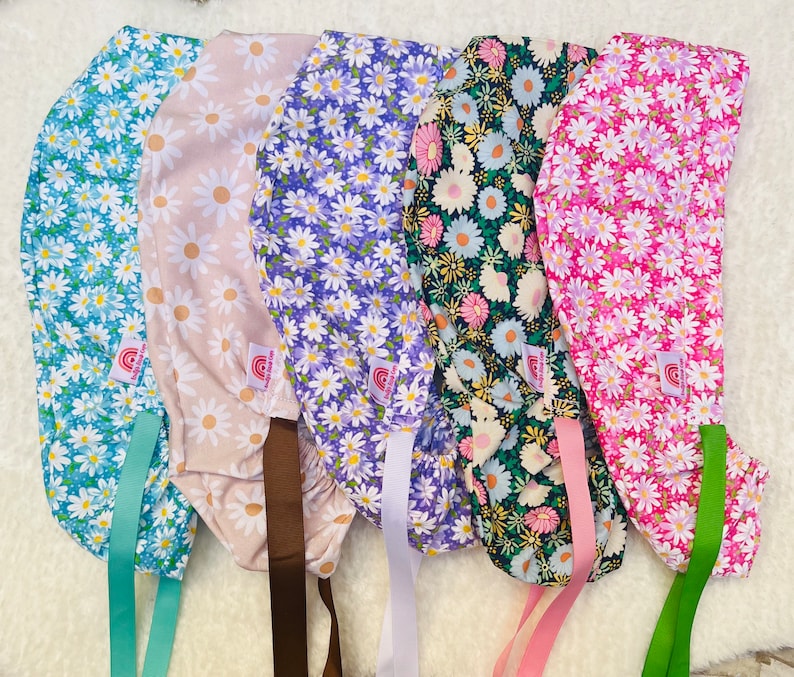 May include: Five colorful scrub hats with floral patterns. The hats are made of fabric and have ties to secure them to the head. The hats are in the following colors: blue, pink, purple, beige, and green.