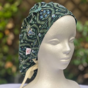 May include: A dark green patterned scrub cap with a floral and butterfly design. The cap features a cream-colored tie in the back and is displayed on a mannequin head. The pattern includes blue, white, and light green accents.