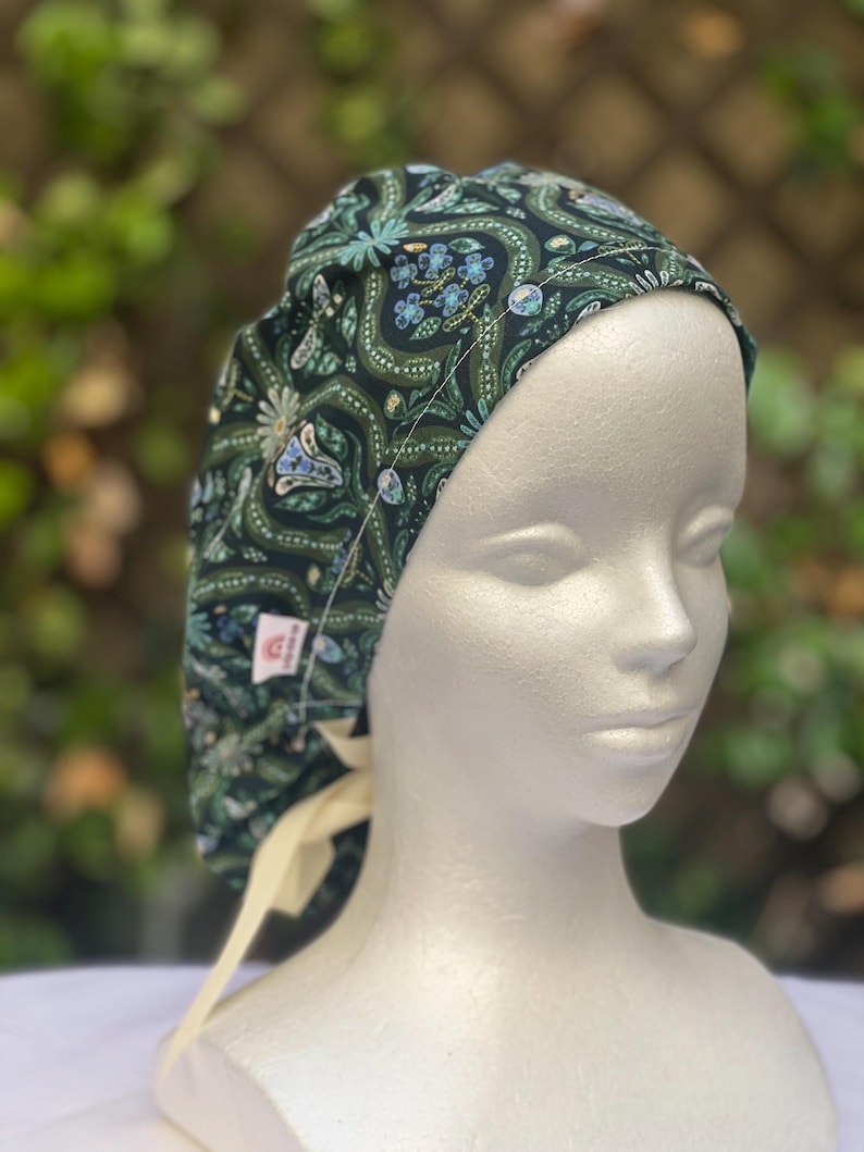 May include: A dark green patterned scrub cap with a floral and butterfly design. The cap features a cream-colored tie in the back and is displayed on a mannequin head. The pattern includes blue, white, and light green accents.
