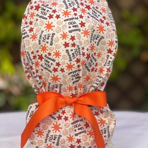 Peut inclure: Un bonnet de chirurgien à motifs avec un nœud orange vif. Le bonnet présente un motif répété d'étoiles rouges, d'éclats de soleil orange et la phrase "Sounds Like a YOU Problem" en texte noir. Le bonnet est fait d'un tissu doux et clair.