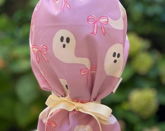 May include: A pink fabric scrub hat with a white ghost and pink bow pattern. The hat is tied with a cream-colored ribbon.
